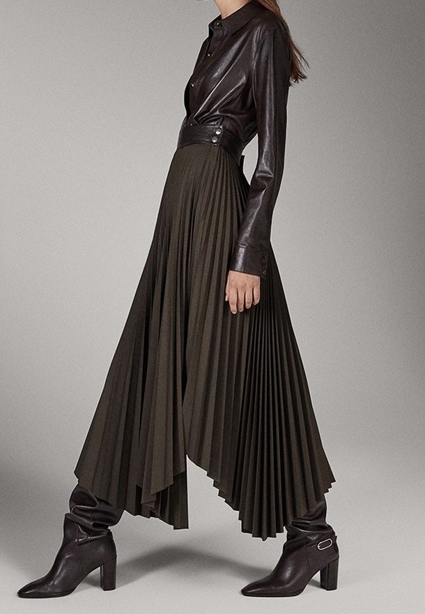 Femme portant une chemise en cuir marron foncé, une jupe plissée asymétrique taille haute et des bottes marron à talons mi-mollet.