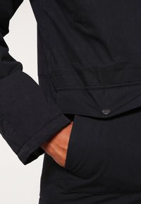 Tmavě modrá bunda s hladkým materiálem, knoflíkovým zapínáním, detaily kapes a prošívanými akcenty na rukávech a lemu.