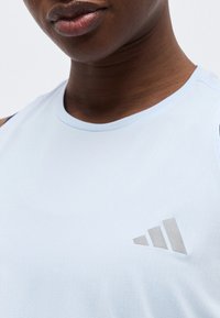 Κοντινό πλάνο σκοτεινόχρωμου ατόμου που φοράει ανοιχτό μπλε αμάνικο τοπ με αντανακλαστικό λογότυπο Adidas στο στήθος.