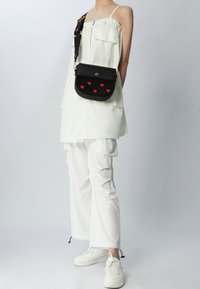 Černá kožená kabelka přes rameno se žlutými srdíčkovými ozdobami, zlatými kovovými prvky a vzorovaným nastavitelným popruhem, v kombinaci s bílým outfitem a teniskami.