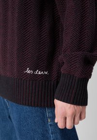 Pleteni pulover s uzorkom riblja kost u tamno burgundiji i crnoj boji, s rebrastim manžetama i obrubom, te bijelim izvezenim logotipom.