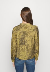 Camicia a maniche lunghe verde oliva con motivo floreale, dotata di un dettaglio a pieghe sul retro e scollo rotondo, indossata con jeans scuri.