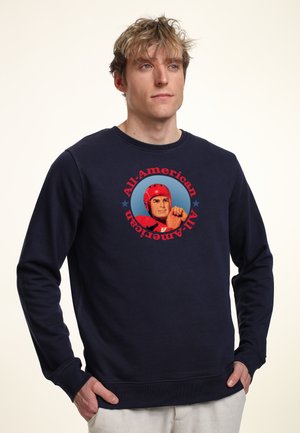 Jeune homme portant un sweat-shirt bleu marine avec un graphisme vintage rouge d'un joueur de football américain et le texte « All-American » dans un cercle sur le devant.