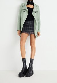 Abrigo verde de mezcla de lana con un diseño de doble botonadura, combinado con un suéter negro corto y una falda mini de cuadros negra. Botas de plataforma negras gruesas.