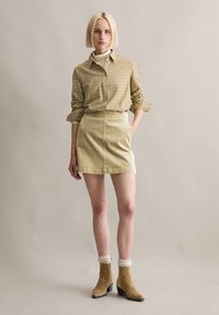 Donna con capelli corti e biondi indossa una camicia beige a quadri abbottonata, una minigonna di velluto a coste chiaro, calzini color crema e stivaletti in camoscio color nocciola, in piedi con espressione neutra.