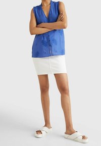 Blouse sans manches bleue avec broderie florale et encolure froncée, associée à une jupe en denim blanche et des sandales à brides croisées blanches.