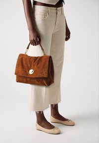 Brown suede handbag with a fold-over flap and gold hardware, held by a person dressed in off-white pants and beige flats with a bow detail.