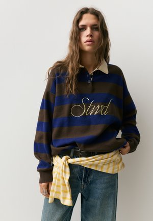 Persoon met lang golvend haar draagt een blauw-bruine gestreepte trui met de tekst "Stndr", een blauwe spijkerbroek en een geel gestreept overhemd om de taille geknoopt.