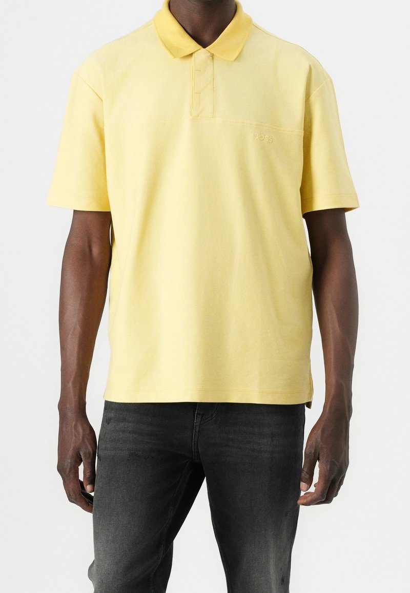 Homme portant un polo jaune à manches courtes avec un col, associé à un jean gris foncé, debout devant un fond uni.