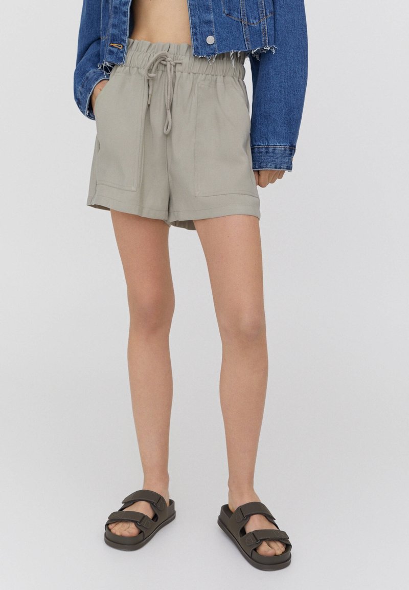 Shorts gris clair avec une ceinture élastique et un cordon de serrage, dotés de deux poches à l'avant, associés à des sandales marron. Veste en denim portée par-dessus.