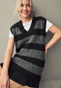 Strikvest med V-hals, der har skiftende sorte og grå striber. Ribbet tekstur, båret over en hvid T-shirt. Casual pasform.