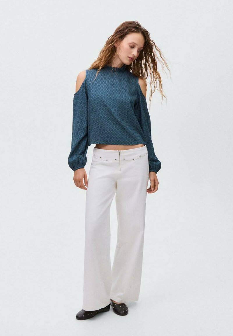 Bluză lungă cu mânecă în teal, cu design cu umeri descoperiți și model texturat, combinată cu pantaloni albi cu talie înaltă și croială largă. Pantofi negri cu puncte.