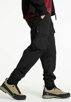 Black cargo pants with multiple pockets, tapered design, and elastic cuffs. Paired with beige and black hiking shoes with lace-up detail.