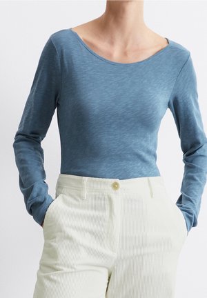 Frau trägt ein enganliegendes blaues Langarmshirt, in weiße, hoch taillierte Cordhosen gesteckt, Hände in den Taschen.