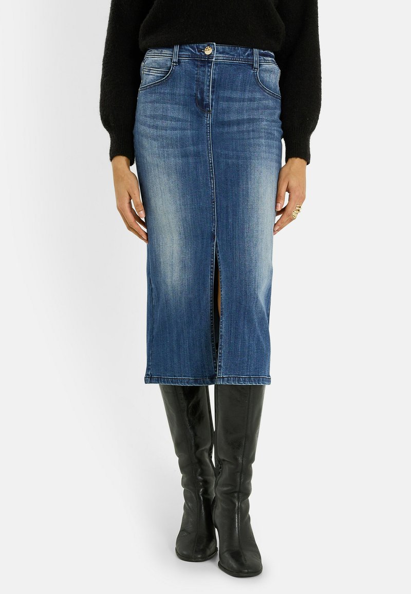 Jupe midi en denim bleu avec fente frontale, portée avec des bottes noires montantes et un pull noir, les mains reposant sur les côtés.