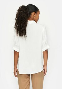 Weiße Bluse mit kurzen Puffärmeln, weicher Textur und lockerer Passform. Verfügt über einen abgerundeten Saum und dezente Raffung an den Ärmelbündchen.