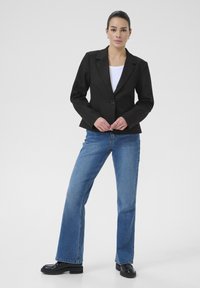 Schwarzer tailierter Blazer mit einem einzigen Knopf, getragen über einem weißen Oberteil. Kombiniert mit hellblauen weiten Jeans und schwarzem Schuhwerk.