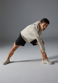 Haut à manches longues gris clair, shorts noirs et baskets claires. Le modèle s'étire, montrant des textures lisses et une coupe détendue sur un fond neutre.