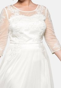 Vitt klänning med genomskinliga långa ärmar, blommig broderi och figursydd bodice. Flödande kjol ger textur och har en slät tygfinish.