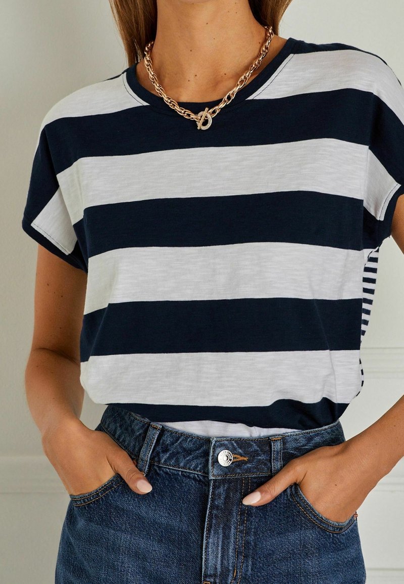 Personne portant un t-shirt rayé bleu marine et blanc rentré dans un jean bleu taille haute, mains dans les poches et un collier en chaîne dorée.
