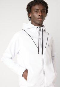 Hoodie blanc zippé avec accents noirs, doté d'une capuche et de poches. Texture lisse, design en couches et logo sur la poitrine.
