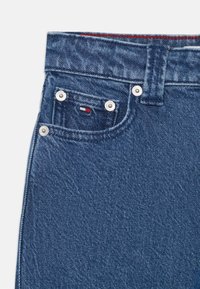 Jean en denim bleu foncé avec un design classique, des accessoires en métal argenté, des détails cousus, et un petit patch logo sur la poche avant.