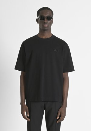 Black short-sleeve t-shirt in cotton, features a crew neck and a subtle logo on the chest. Worn with black tailored trousers and sunglasses.