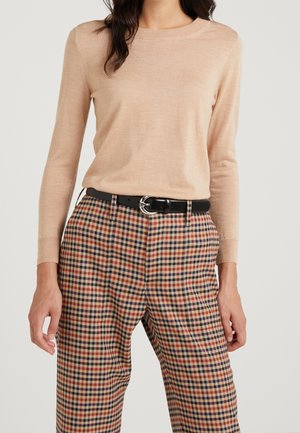 Femme portant un pull beige à manches longues rentré dans un pantalon à carreaux taille haute avec une ceinture noire à boucle argentée.
