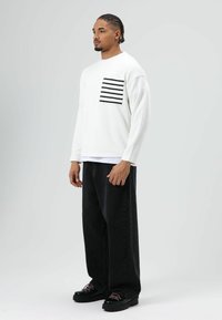 Witte blouse met lange mouwen en een zwart gestreepte zak, gecombineerd met zwarte broek met wijde pijpen en stevige zwarte schoenen met roze accenten.