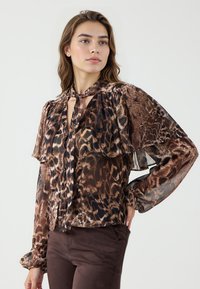Blouse à imprimé animal marron avec des manches longues, un col à nouer et des volants superposés. Fabriquée en tissu léger et transparent.