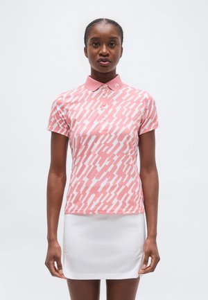 Kvinde iført en pink og hvid mønstret polo-shirt med krave og en ensfarvet hvid nederdel, stående mod en hvid baggrund.