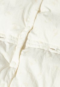 Cremefarbige, gesteppte Bettwäsche mit einem dezenten Muster, versehen mit runden weißen Knöpfen entlang der Naht und einer glatten, weichen Textur.