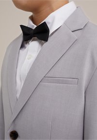 Ljusgrå kavaj med snedskuren slag, enkel knappstängning och ficka med flik. Matchad med en vit skjorta och svart fluga.