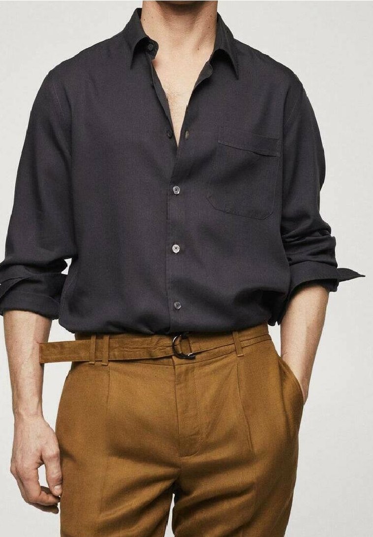 Homme portant une chemise à boutons gris foncé avec les manches retroussées et un pantalon taille haute marron moutarde avec une ceinture assortie.