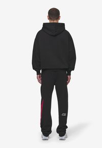 Černá mikina s kapucí a uvolněným střihem, v kombinaci s černými tepláky s růžovým grafickým motivem na levé noze.