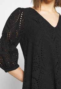 Mustat mekko, jossa on V-pääntie, upeita leikattuja kuvioita, puffeja hihoja ja teksturoidusta kankaasta. Huomattavia kukkamotiiveja on näkyvissä.