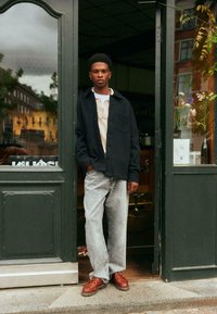 Black button-up shirt over a beige textured sweater, paired with light gray jeans and brown lace-up shoes, standing in a doorway.