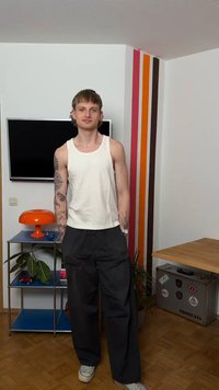 Νέος άνδρας με τατουάζ, φορώντας λευκό φανελάκι και μαύρο παντελόνι, στέκεται σε ένα δωμάτιο με πολύχρωμο ριγέ τοίχο, πορτοκαλί λάμπα και μεταλλική μονάδα ραφιών.