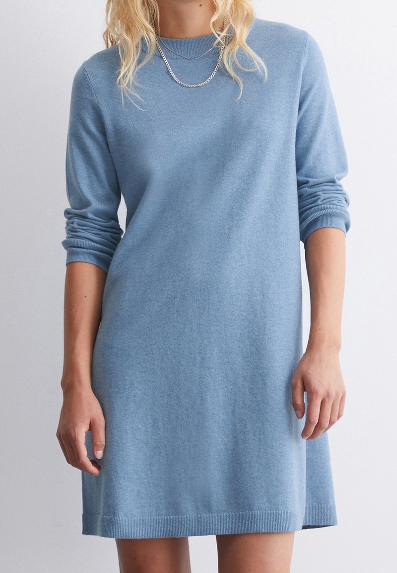 Femme portant une robe pull en maille bleu clair à manches longues avec un collier chaîne argenté, se tenant devant un fond blanc uni.