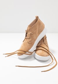 Baskets en daim brun clair avec semelles en caoutchouc blanches, présentant un design à lacets et des lacets plats tombant librement à l'avant. Surface supérieure texturée.