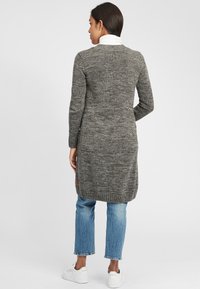 Long grey knitted cardigan with a textured pattern, ribbed cuffs, and a side logo tag, worn over blue jeans and white sneakers.