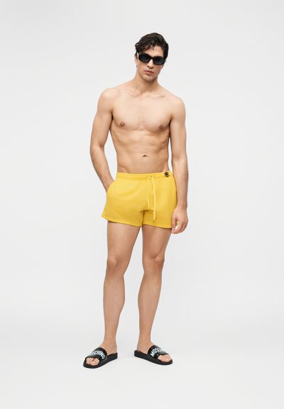Άνδρας χωρίς πουκάμισο, φορώντας κίτρινα μαγιό σορτς, μαύρα γυαλιά ηλίου και μαύρα σανδάλια slide με όνομα μάρκας, στέκεται μπροστά σε απλό φωτεινό φόντο.