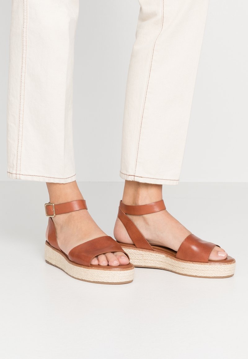 Pieds portant des sandales plateformes en cuir marron à bride cheville associées à un pantalon beige clair raccourci, sur un fond uni.