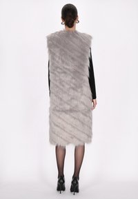 Chaleco de piel sintética gris con forma larga y recta, que presenta una superficie texturizada y un patrón en capas; modelo combinado con una blusa negra y zapatos.