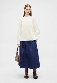 Ελαφρύ μπεζ πουλόβερ πλεκτό με ριμπ τελείωμα, σε συνδυασμό με μια μακριά σκουρομπλέ φούστα από τζιν. Αξεσουάρ περιλαμβάνουν καφέ τσάντα και μποτάκια με τακούνι.