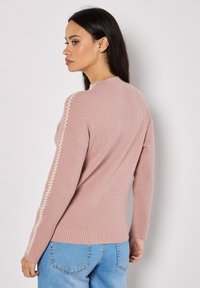Rippensweater in Rosa mit langen Ärmeln, hohem Kragen und dekorativer cremefarbener Naht entlang der Schulter. Kombiniert mit blauen Jeans.