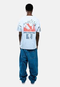 Szara bawełniana koszulka z wielokolorową grafiką z tyłu, zawierającą tekst i wzór płomienia, zestawiona z luźnymi niebieskimi dżinsami.