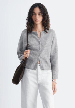 Femme aux cheveux bouclés portant un cardigan gris boutonné et un pantalon blanc, tenant un sac à épaule foncé, sur un fond clair uni.