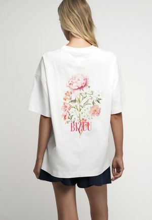 Kobieta z długimi blond włosami nosi białą oversize koszulkę z różowo-białym kwiatowym wzorem i tekstem "BIZUU" na plecach.