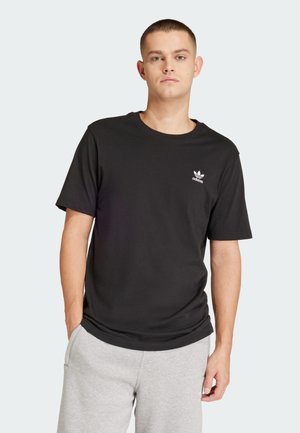 Černé tričko s krátkým rukávem vyrobené z bavlny, s bílým logem Adidas na levém hrudníku. Stylizováno s šedými tepláky.